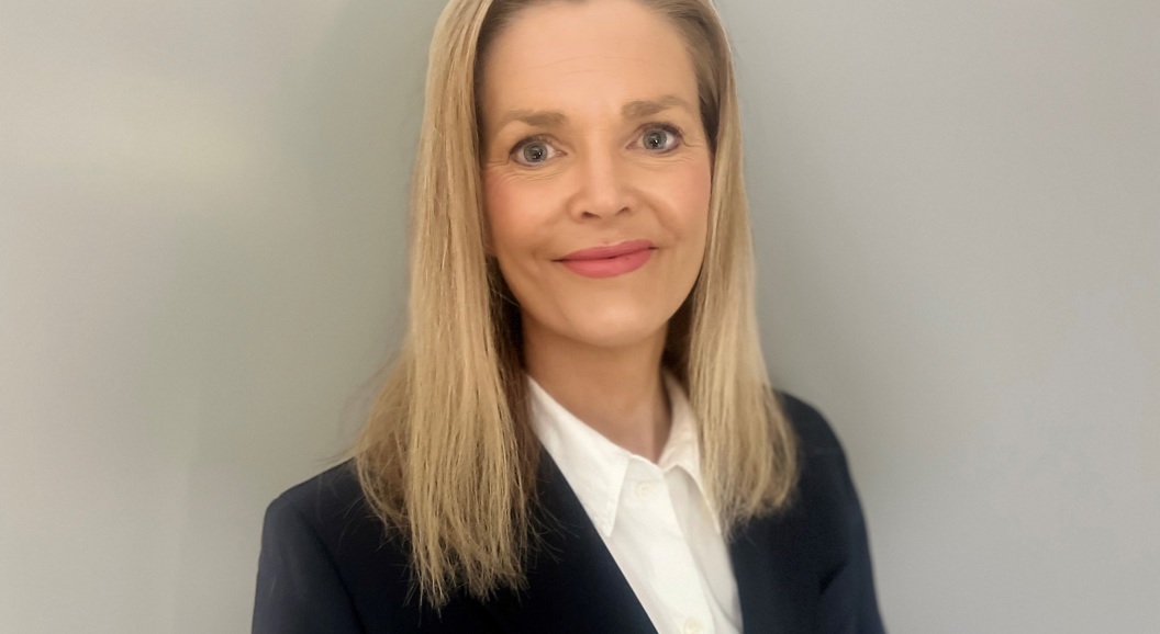 Kvinne med langt blondt hår iført ein mørk dressjakke og kvit skjorte, framfor ein grå bakgrunn.