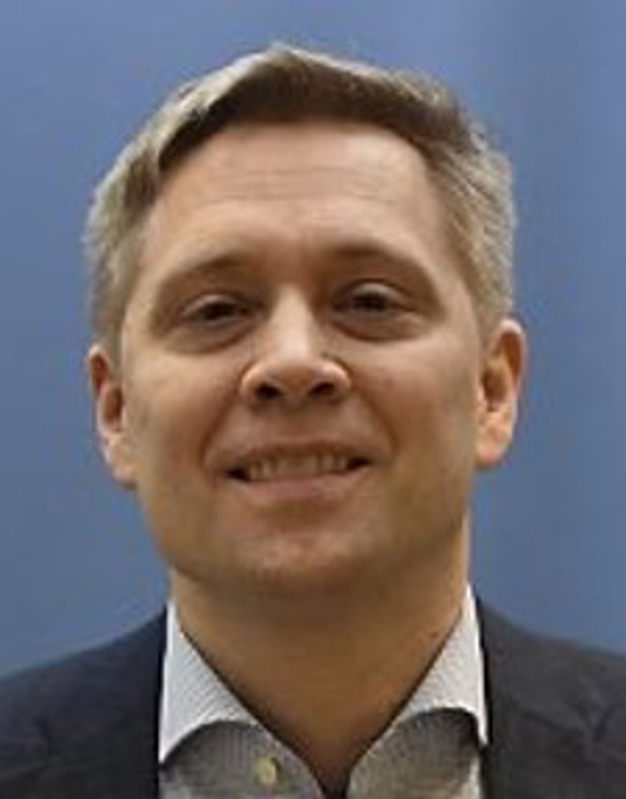 Ein person med kort, grått hår og mørk dress står framfor ein blå bakgrunn, ser rett fram og smilar