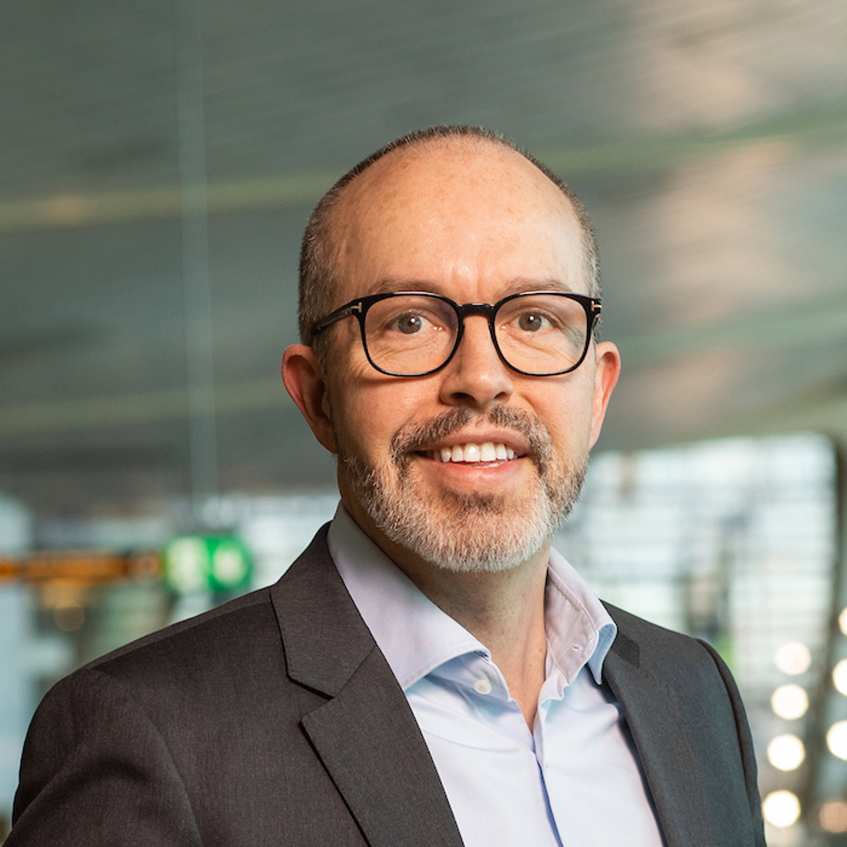 Ein mann med briller og skjegg i formell dressjakke smiler, med ei moderne kontorinnreiing i bakgrunnen.