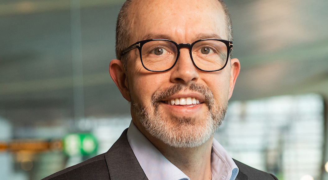 Ein mann med briller og skjegg i formell dressjakke smiler, med ei moderne kontorinnreiing i bakgrunnen.