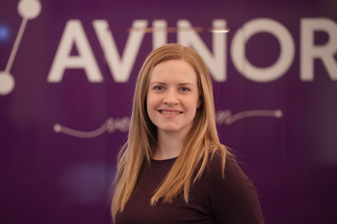 Kvinne med langt blondt hår smiler framfor ein lilla bakgrunn med teksten "AVINOR".