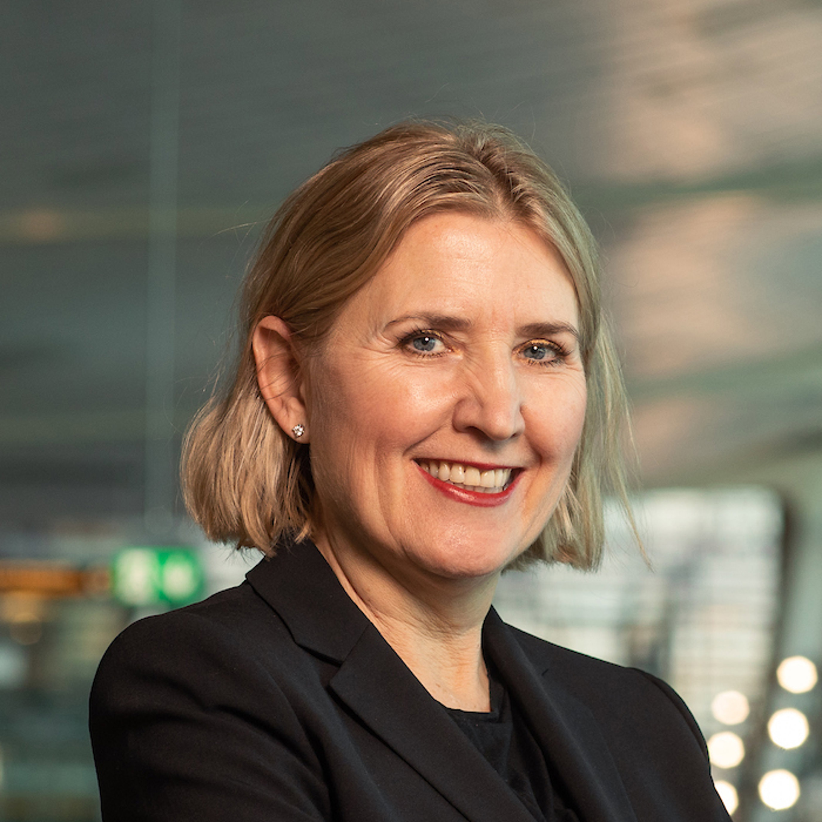 Kvinne med skulderlangt blondt hår og svart blazer smiler i eit moderne kontormiljø.