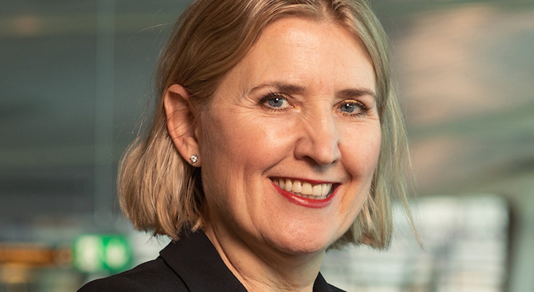 Kvinne med skulderlangt blondt hår og svart blazer smiler i eit moderne kontormiljø.