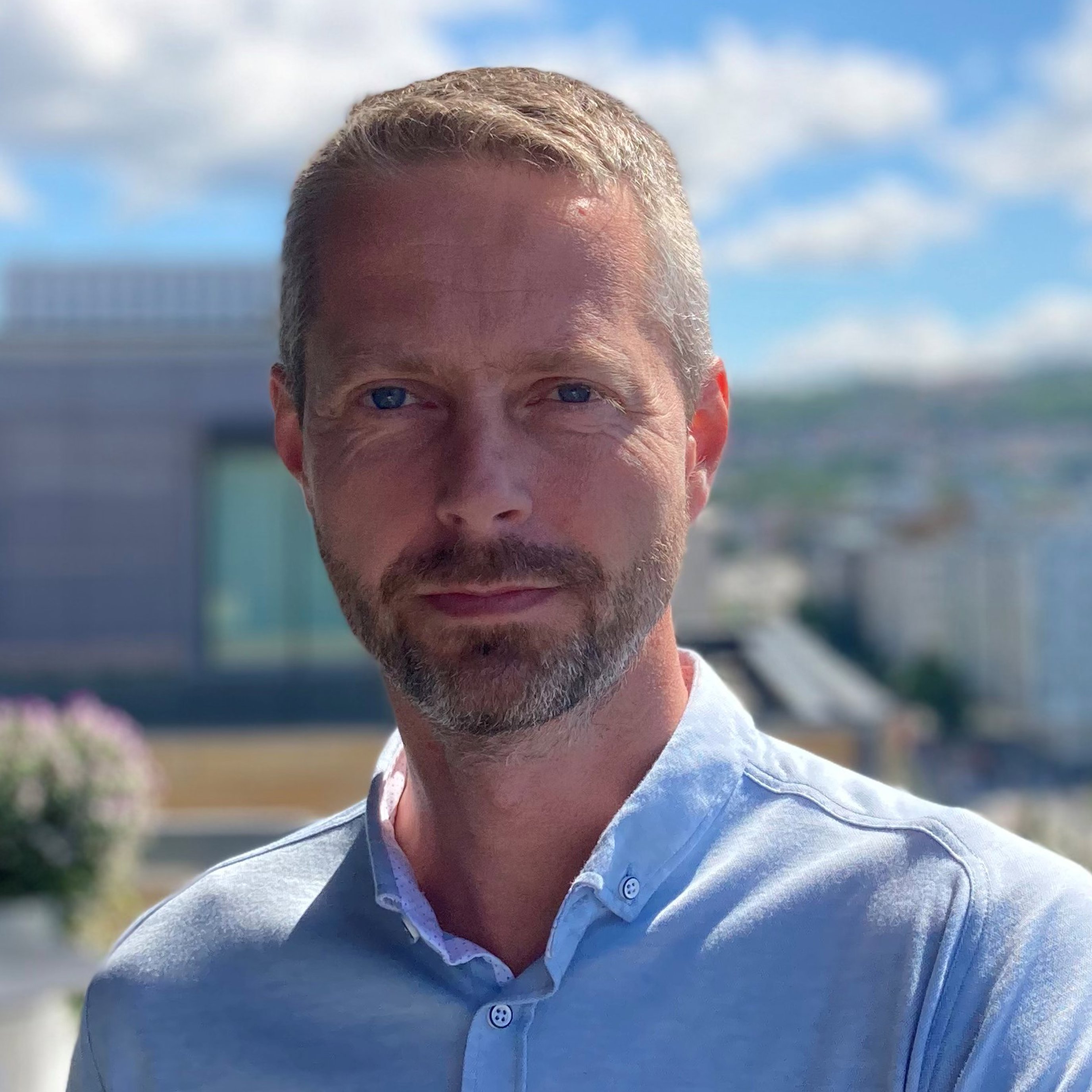 Mann i utendørs omgivelser iført en lyseblå skjorte, med en uskarp bakgrunn av et bylandskap og overskyet himmel