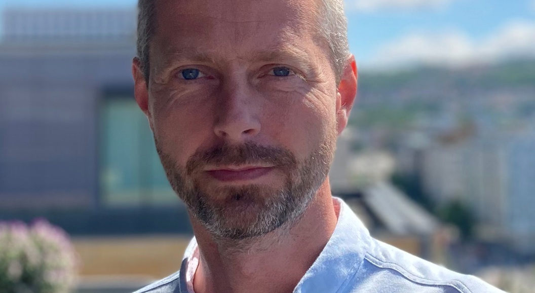 Ein mann med kort hår og skjegg, iført ein lyseblå skjorte, står utandørs med uklårt bylandskap i bakgrunnen. Blå himmel og skyer synes over han