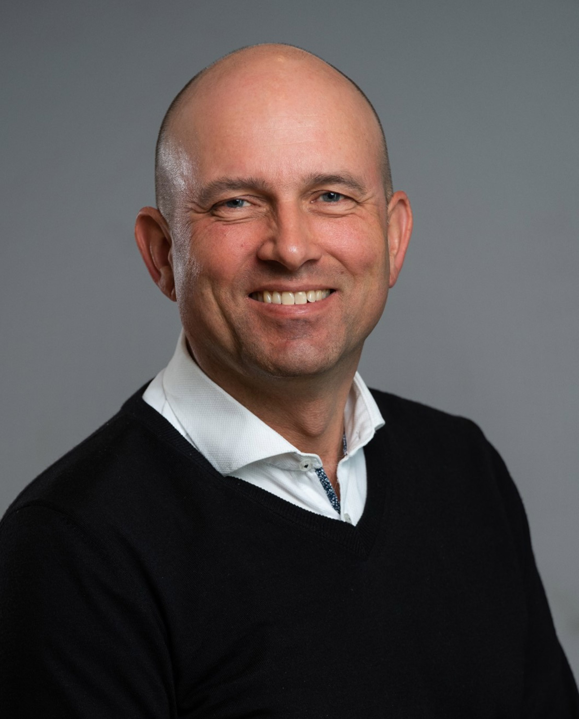 Ein mann med skalla hovud, smiler iført kvit skjorte og svart genser mot ein grå bakgrunn
