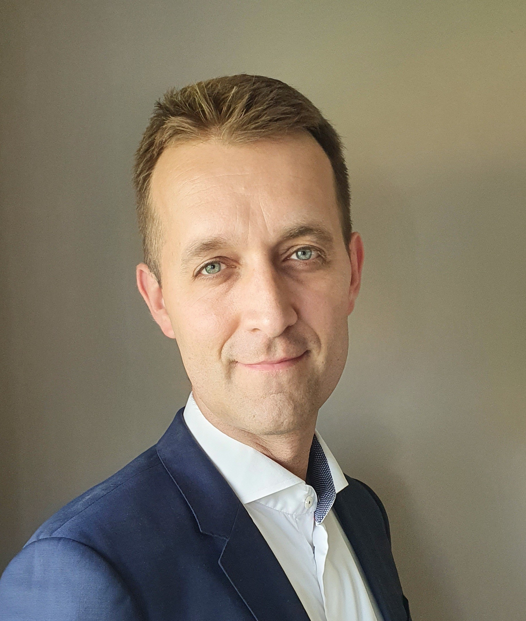 A man in a blazer smiles at the camera, with a neutral background.
