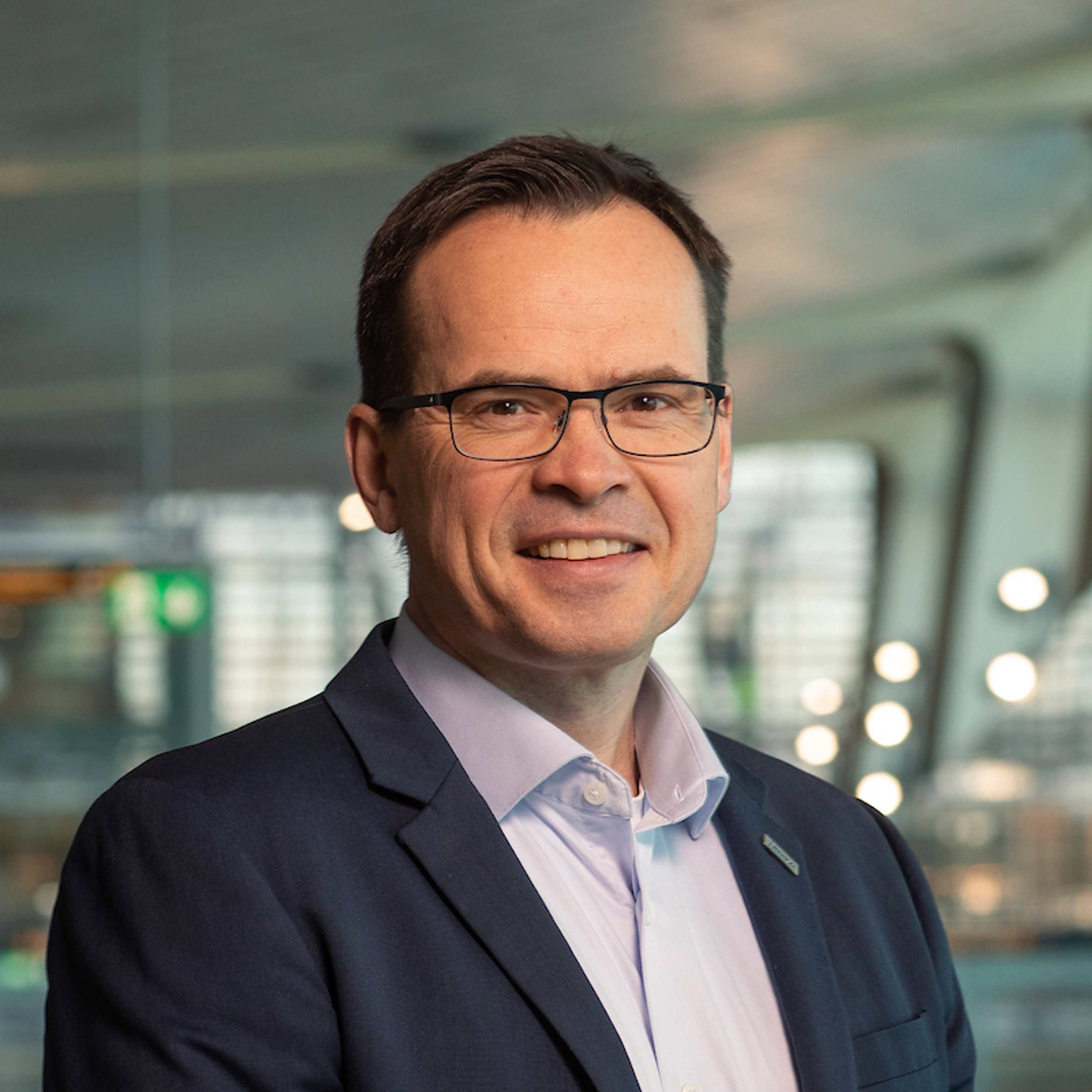 Forretningsmann i dress og briller står innendørs med moderne arkitektur i bakgrunnen, lyssatt med mykt naturlig lys.