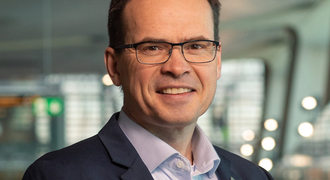 Forretningsmann i dress og briller står innandørs med moderne arkitektur i bakgrunnen, lyssatt med mjukt naturleg lys.