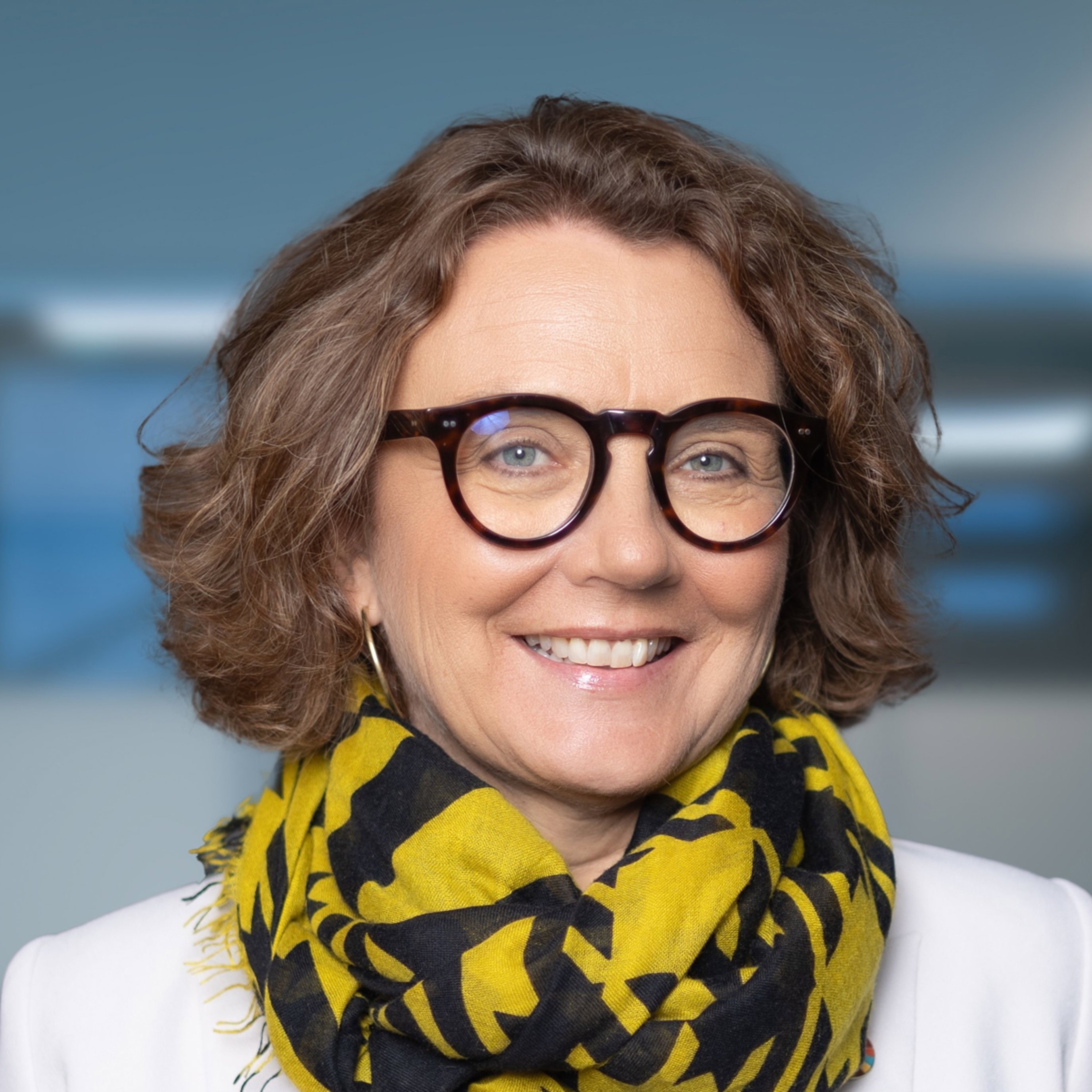 Kvinne med briller og krøllete hår smiler mot kameraet, iført en gul og svart mønstret skjerf og en hvit jakke, i et moderne innendørsmiljø.