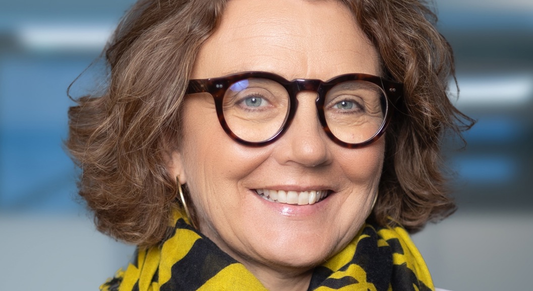 Kvinne med briller og krøllete hår smiler mot kameraet, iført eit gult og svart mønstra skjerf og ei kvit jakke, i eit moderne inneområde.