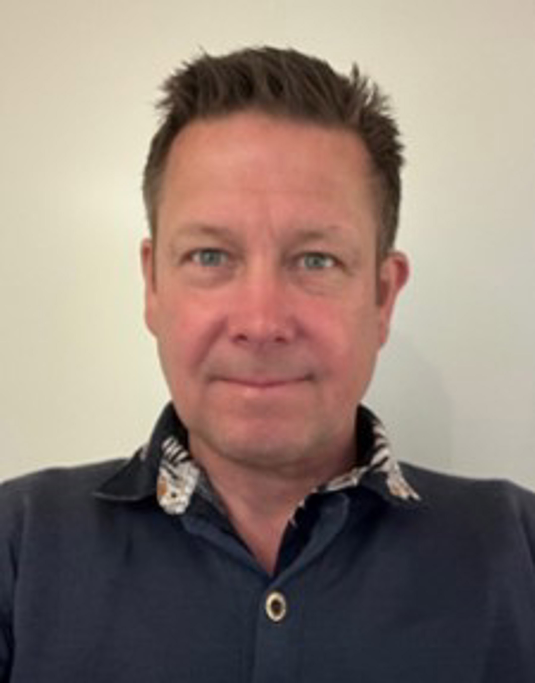 Mann i blå skjorte med kort brunt hår, smiler svakt mot kameraet framfor kvit bakgrunn