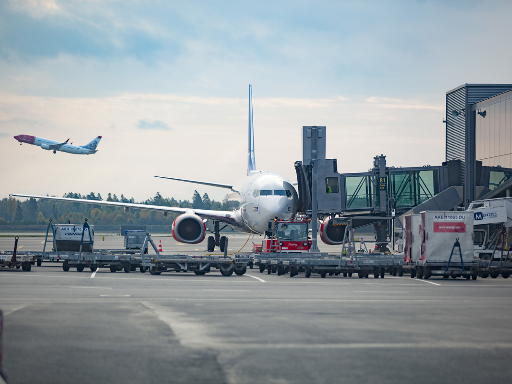 Flyplass gate med fly ved terminal, parkert under flygebro, medan eit anna fly tek av i bakgrunnen, over overskya himmel