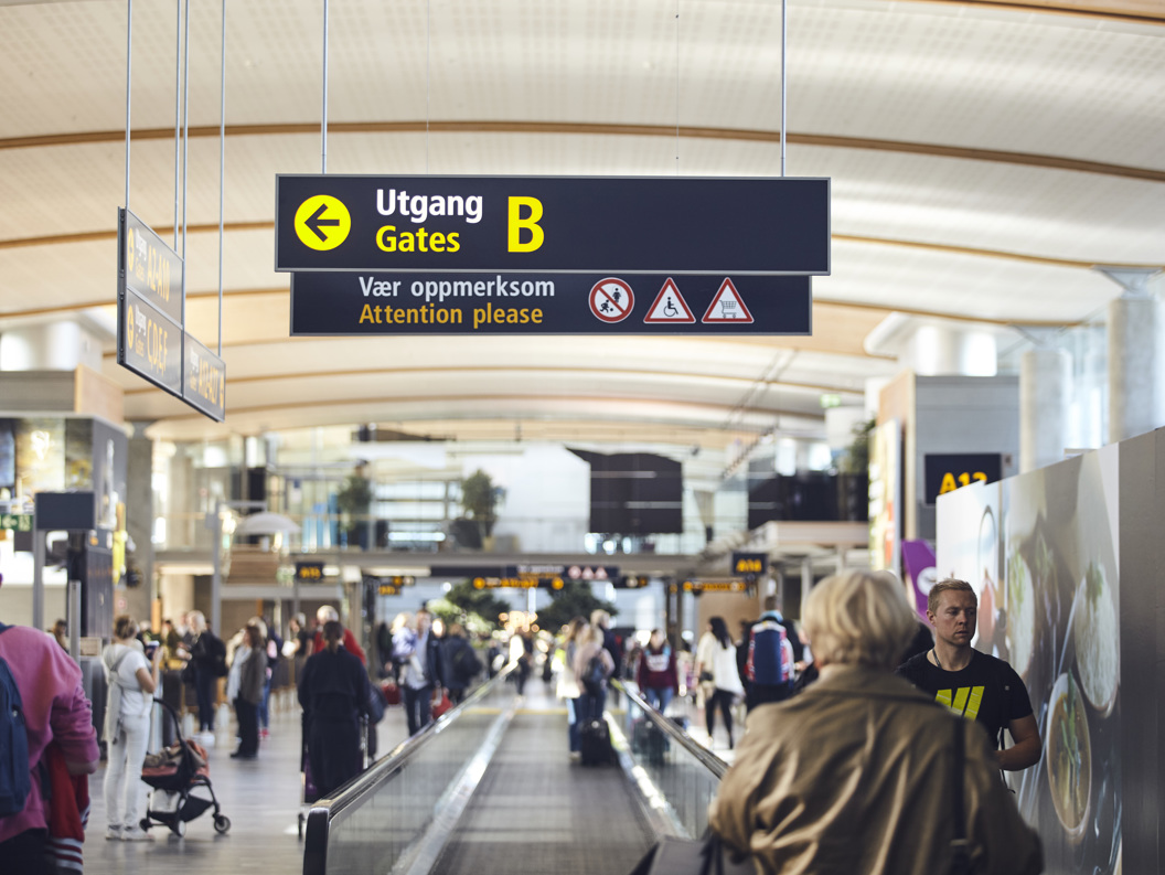 Folk på flyplass beveger seg mot utgang B, med skilt som viser veg og gir åtvaringar om røyking, mobilbruk, og rullestoltilgjenge.