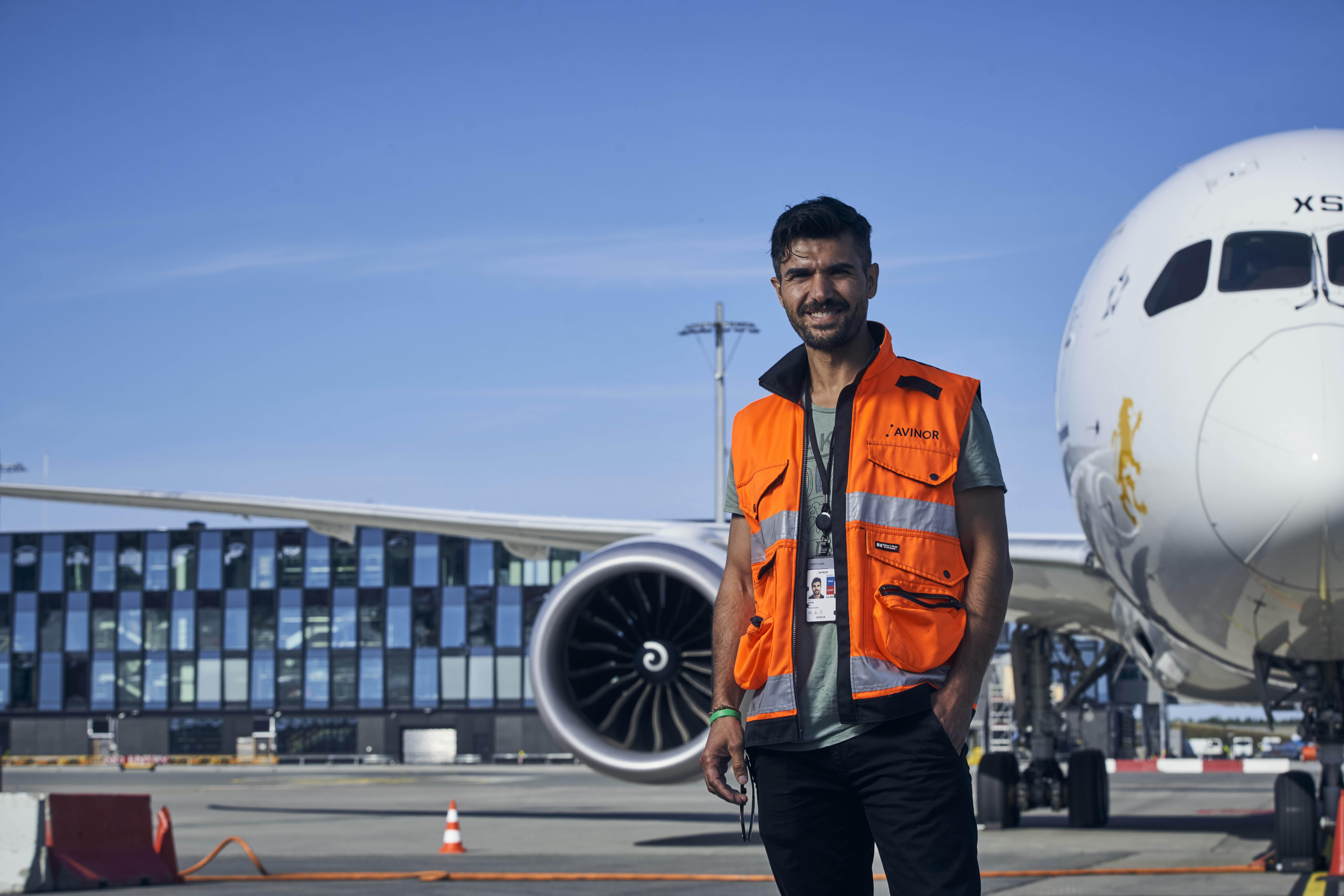 Ein mann i oransje tryggleiksvest står framfor eit fly på ein flyplass, med moderne terminalbygg i bakgrunnen
