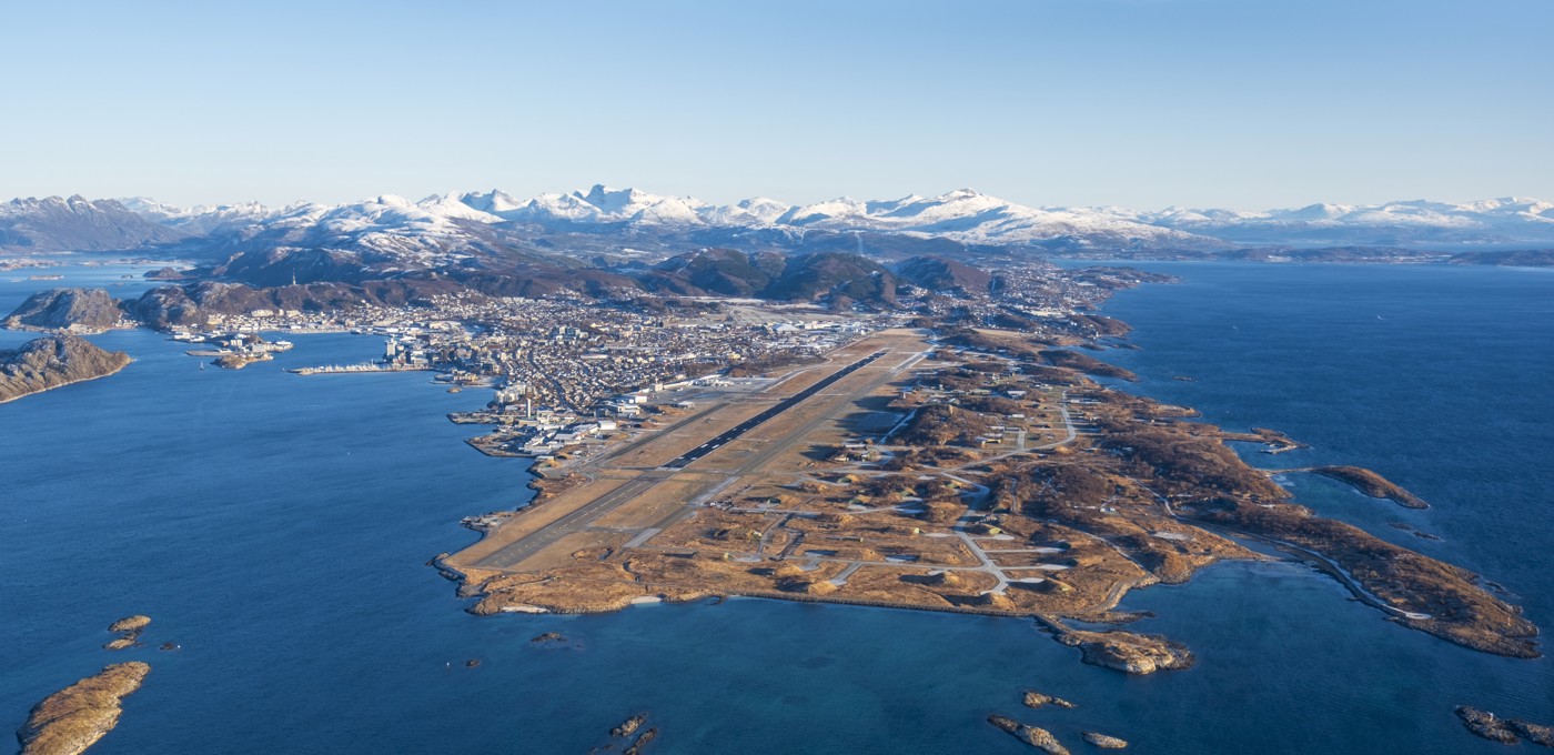 Luftfoto av Bodø i Norge med Bodø lufthavn i forgrunnen og snødekte fjell i bakgrunnen. Byen ligger langs kystlinjen med det blå havet på begge sider av halvøya.
