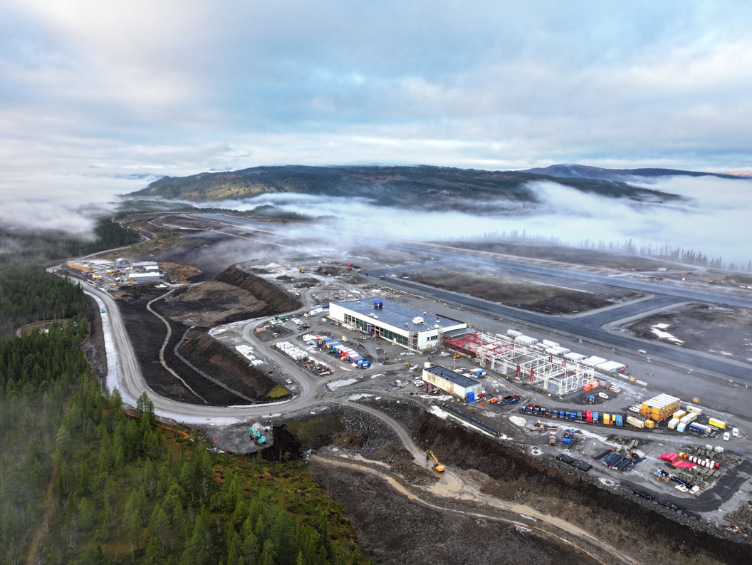Luftfoto av en industriell anleggsplass i et kupert landskap med tåke over skogen, viser bygg og kjøretøy på området