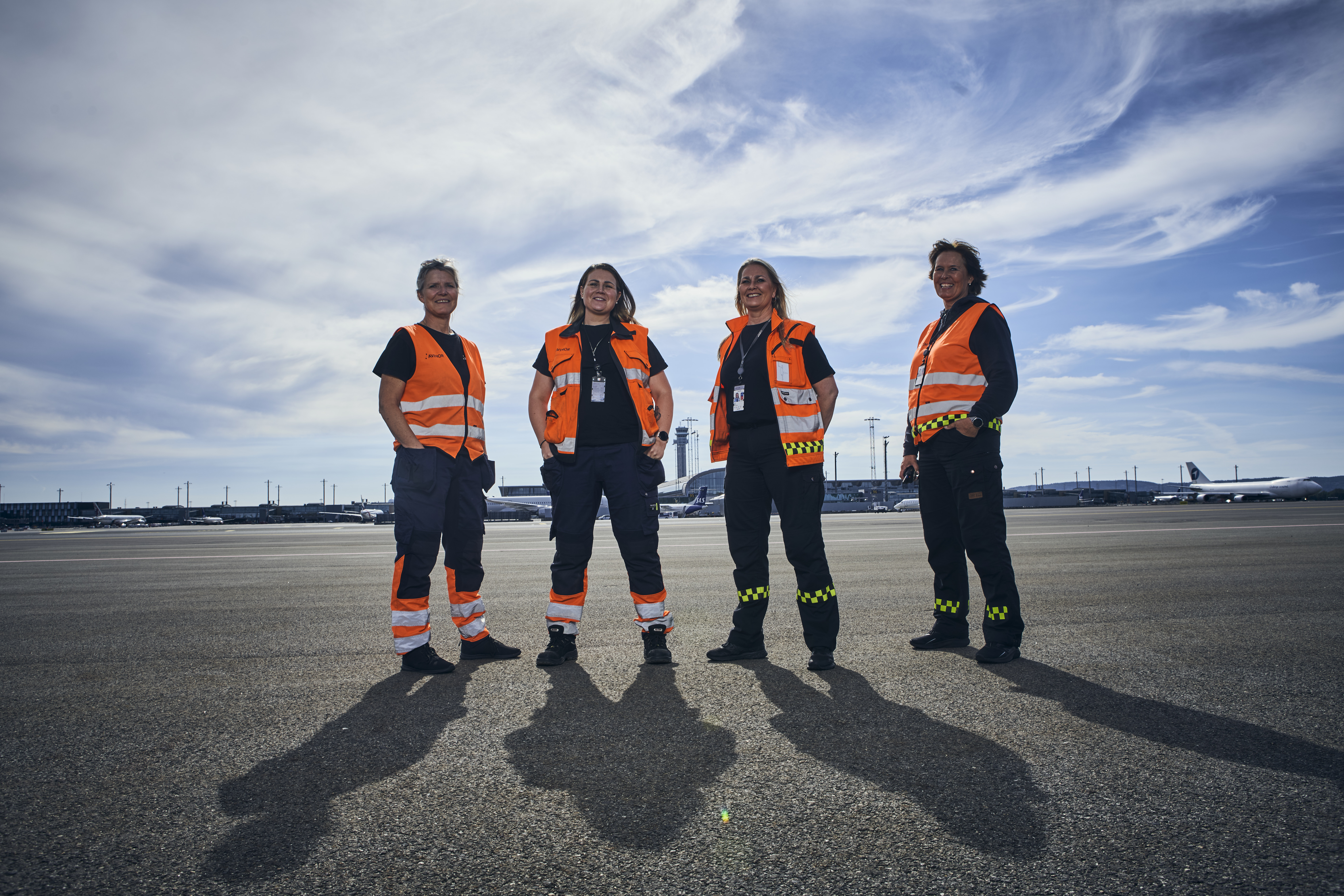 Four airport ground crew members wearing orange safety vests standing on a tarmac with airplanes and control towers in the background under a cloudy sky