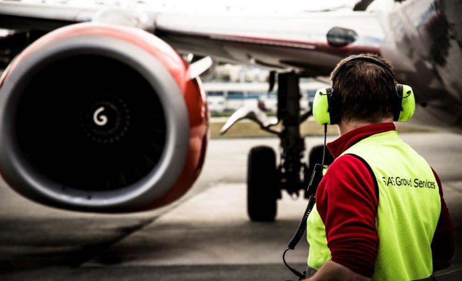 Flyplasspersonale med høyrselsvern overvaker behandlinga av eit fly på bakken framfor ein jetmotor.