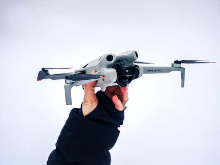 Hånd som holder en grå drone med fire propeller mot en hvit bakgrunn.