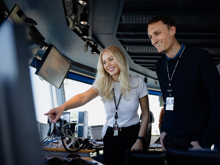 To personar i eit kontrollrom som ser på ein skjerm, smilande og engasjerte i arbeidet sitt.