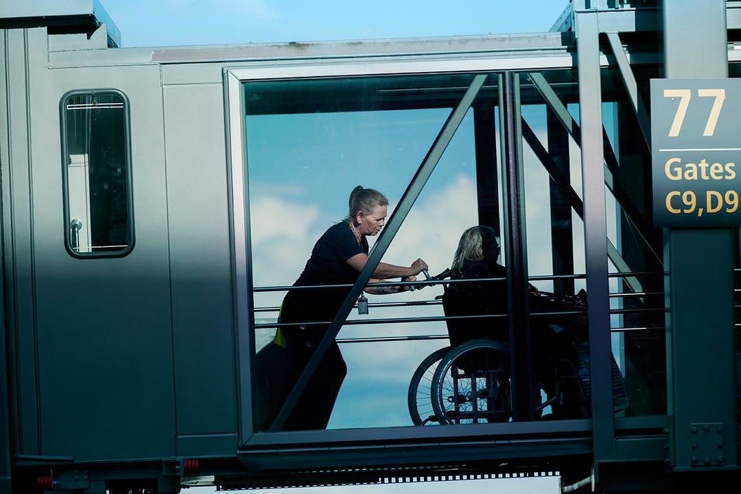 Ein tilsett hjelper ein person i rullestol gjennom ein gangbru på ein flyplass. Skilt til port 77 med portnummer C9 og D9 er synleg