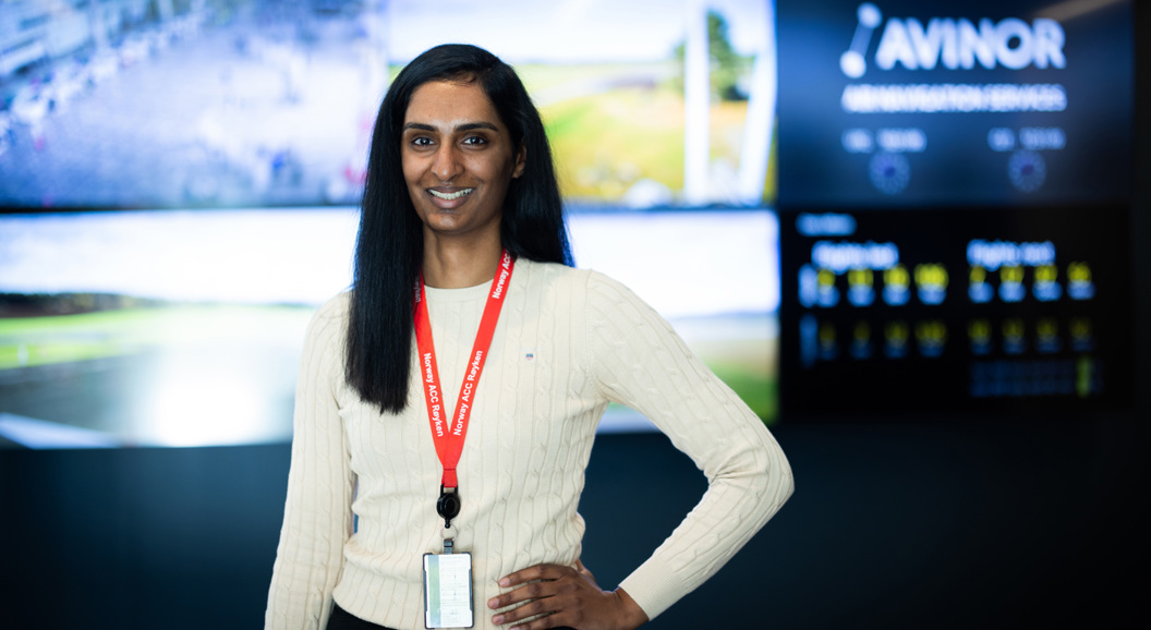 Kvinne med langt mørkt hår står smilende foran flere skjermer med Avinor-logoen i bakgrunnen, iført hvit genser og rød ID-lanyard.