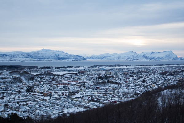 Vinterlandskap i Norge med snødekte fjell, hus, og en fjord i bakgrunnen under en overskyet himmel.