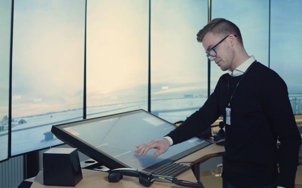 Mann jobber ved en stor berøringsskjerm på en flykontrollstasjon, med panoramautsikt over flyplassen i bakgrunnen.