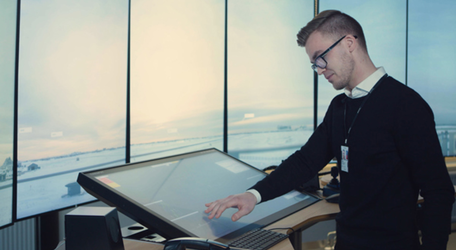 A man works at a large touch screen at an air traffic control station, with a panoramic view of the airport in the background.