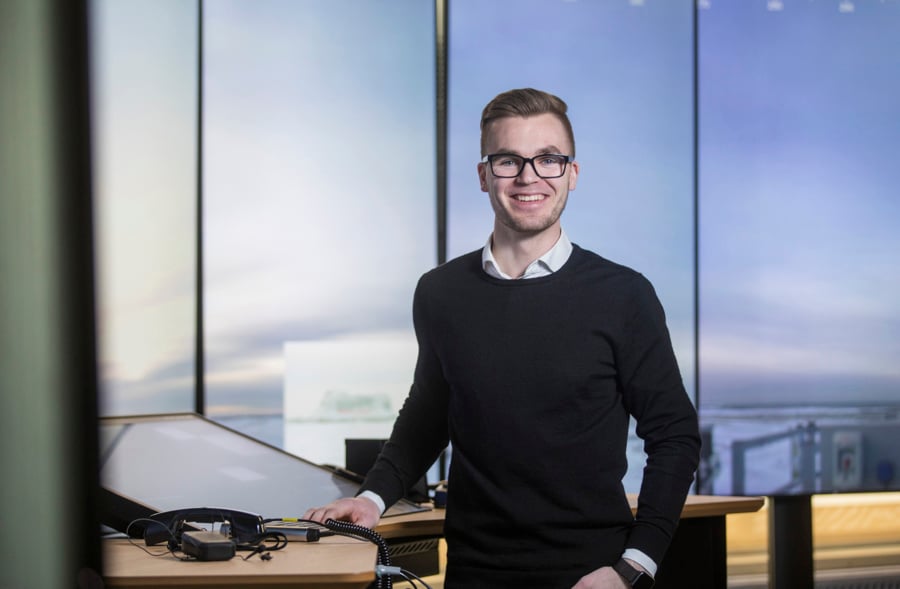 A smiling person with glasses stands at a desk with technology equipment, in a modern office setting.