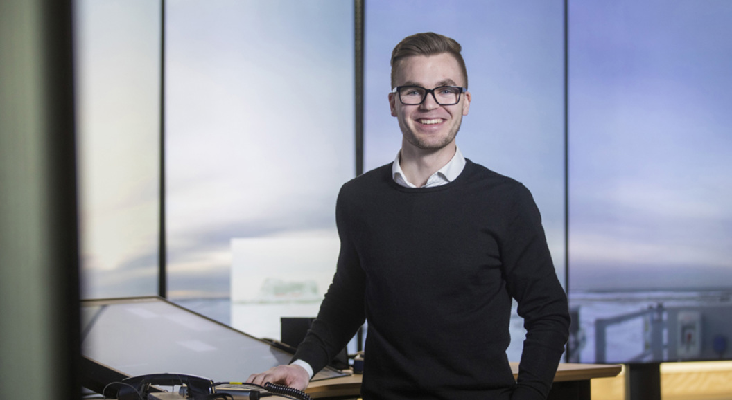 Ein smilande person med briller står ved eit skrivebord med teknologiutstyr, mot ei moderne kontorinnstilling.