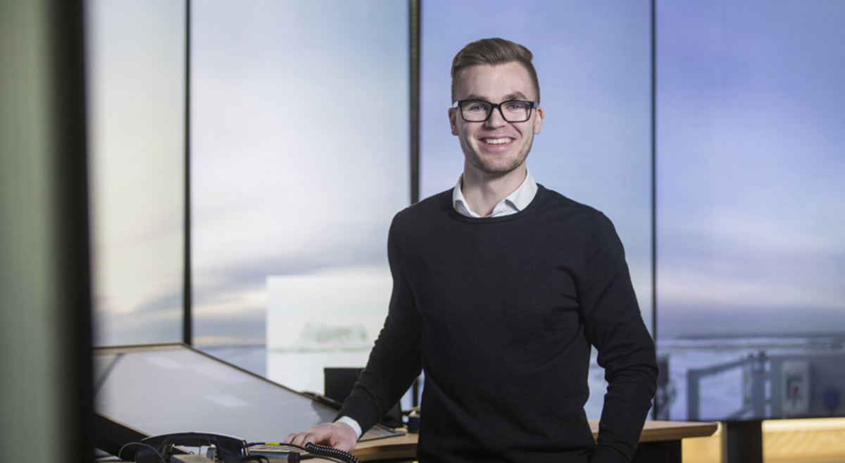 A smiling person with glasses stands at a desk with technology equipment, in a modern office setting.