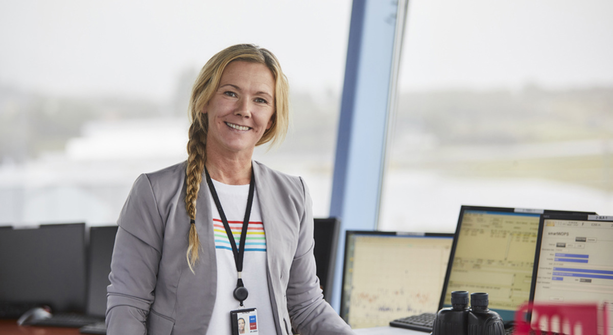 Woman in control room with computer screens in the background, wearing a gray jacket and an ID card.