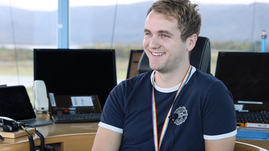A young man is sitting smiling at a modern workspace with multiple computer screens in the background, wearing a dark blue t-shirt and a rainbow-colored lanyard. The background shows a landscape through the window.