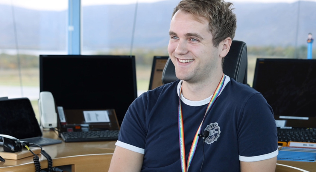 Ein ung mann sit smilande ved eit moderne arbeidsområde med fleire dataskjermer i bakgrunnen, iført ei mørkeblå t-skjorte og eit regnbogefarga lanyard. Bakgrunnen viser eit landskap gjennom vindauget.