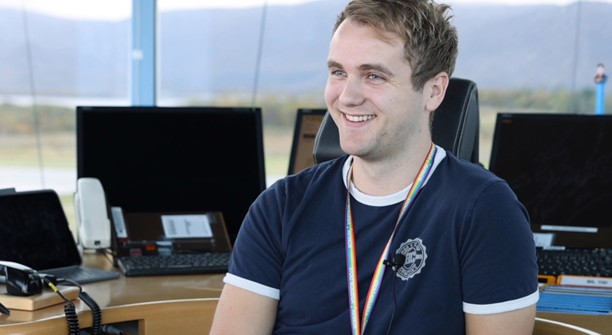 A young man is sitting smiling at a modern workspace with multiple computer screens in the background, wearing a dark blue t-shirt and a rainbow-colored lanyard. The background shows a landscape through the window.