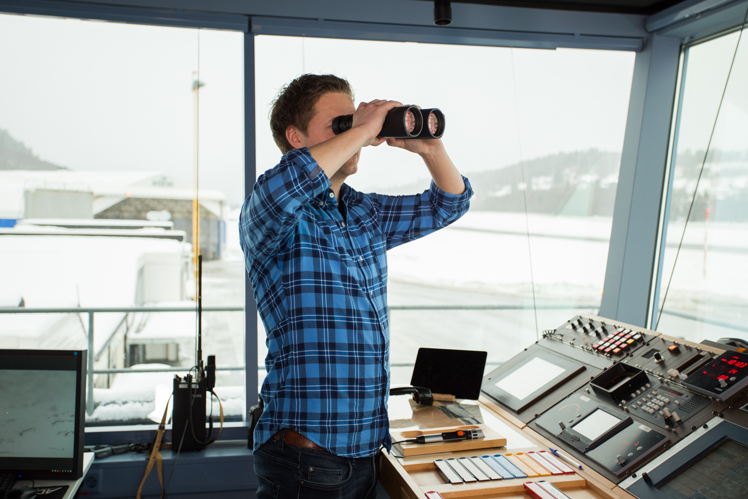 Man in a blue checkered shirt uses binoculars in a control room with multiple screens and control panels. View of snow-covered landscape through the windows.