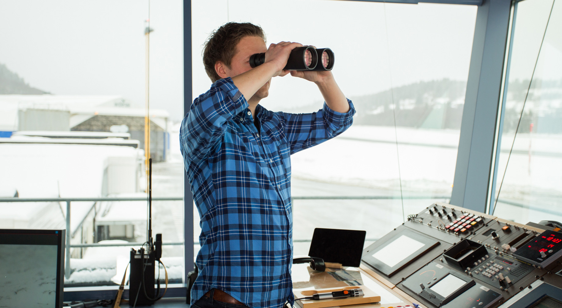 Mann i blå rutete skjorte bruker kikkert i et kontrollrom med flere skjermer og kontrollpaneler. Utsikt over snødekket landskap gjennom vinduene.