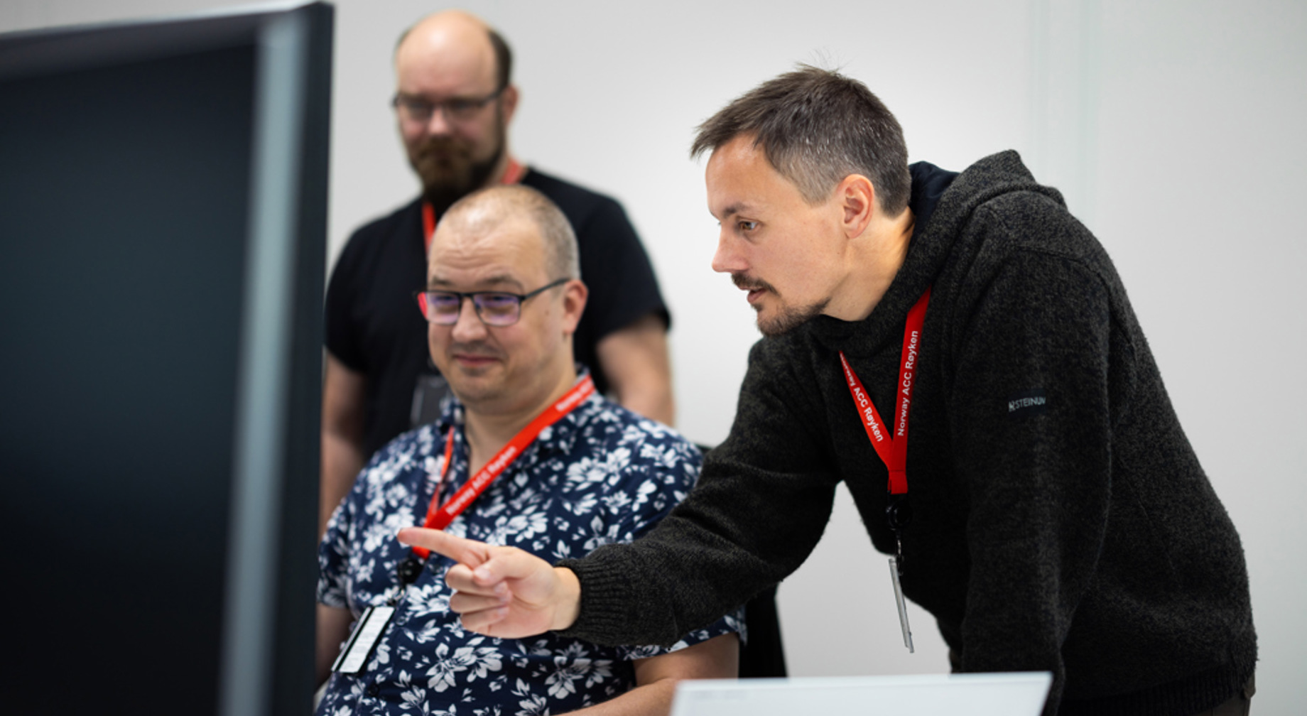 Tre menn samarbeider på et kontor, med en som peker på en dataskjerm mens de andre observerer oppmerksomt