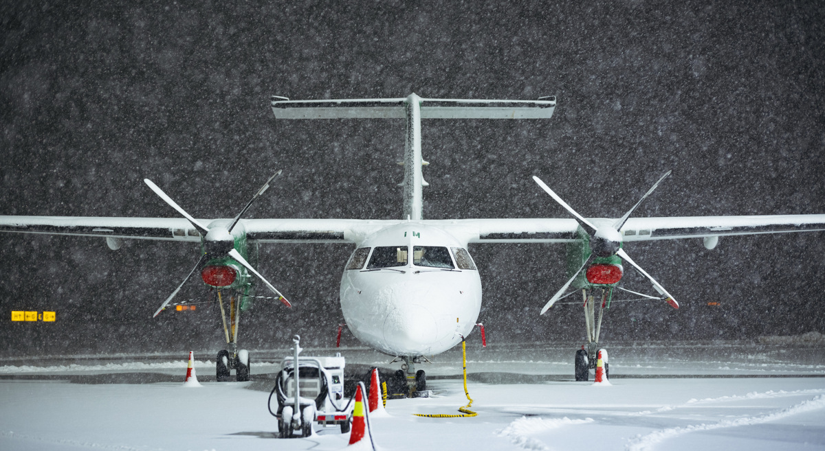 Turbopropfly stående på snødekt flyplass under kraftig snøfall, sett forfra med to propeller og opplyst neseparti