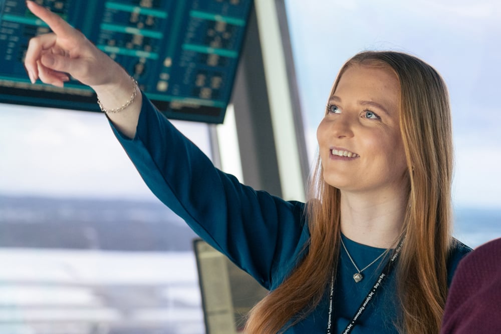 Kvinne peker mot skjerm i kontrollrom, smiler og ser fokusert ut. Skjermer med data i bakgrunnen.