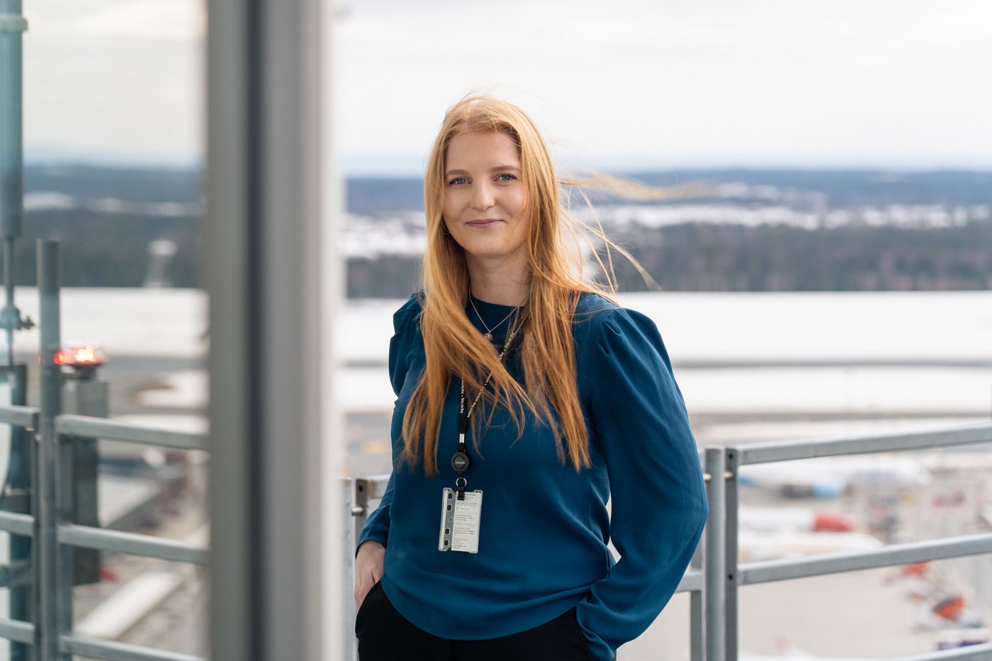 Kvinne med langt rødt hår står utendørs med en flyplass i bakgrunnen, iført en blå genser og ID-kort rundt halsen.