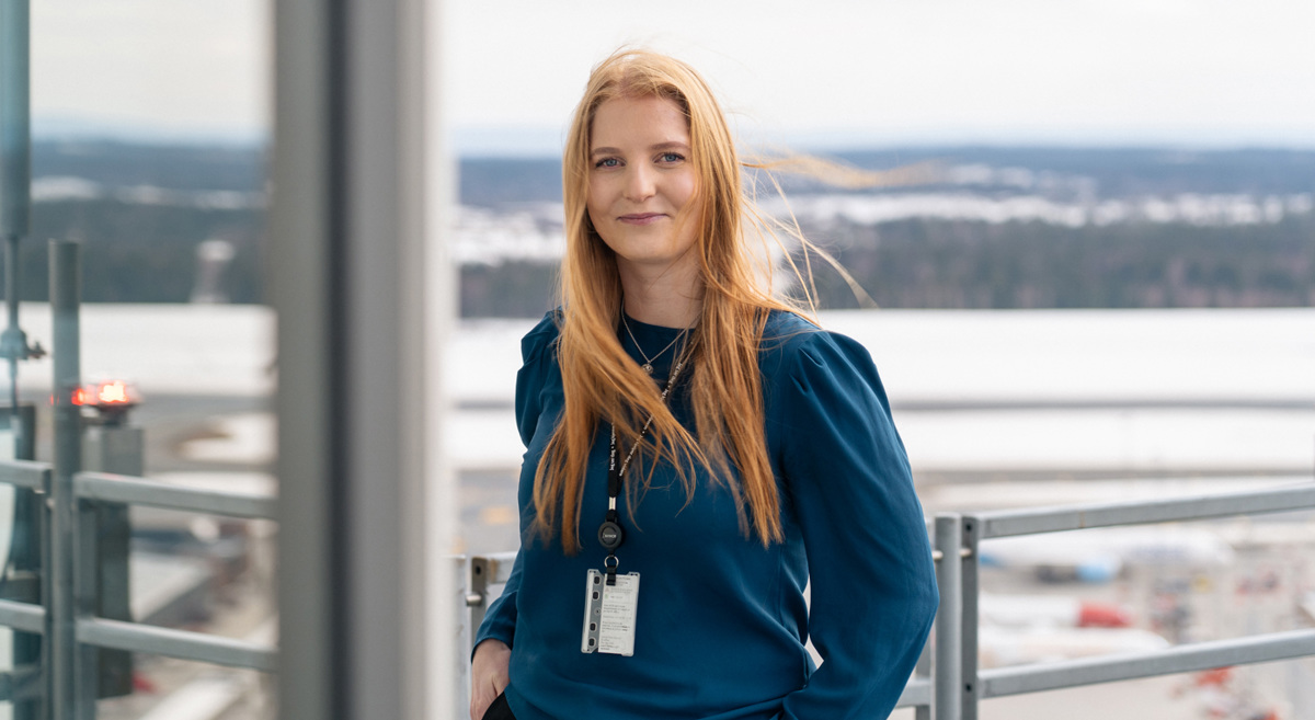 Kvinne med langt rødt hår står utendørs med en flyplass i bakgrunnen, iført en blå genser og ID-kort rundt halsen.