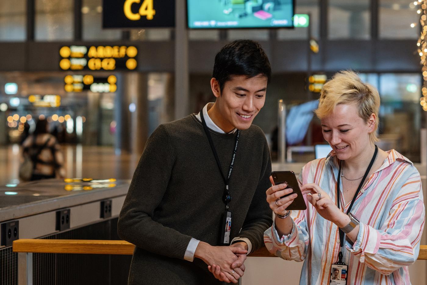 To personer ser på en mobiltelefon på en moderne flyplass, med skilt og informasjonstavler i bakgrunnen.