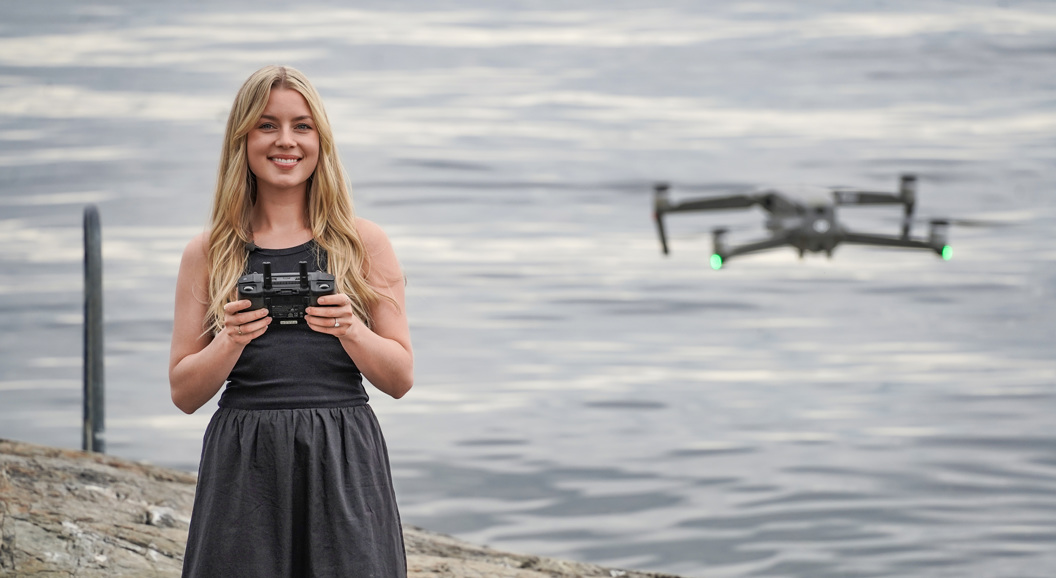 Smilende kvinne styrer en drone med fjernkontroll ved vannet\
