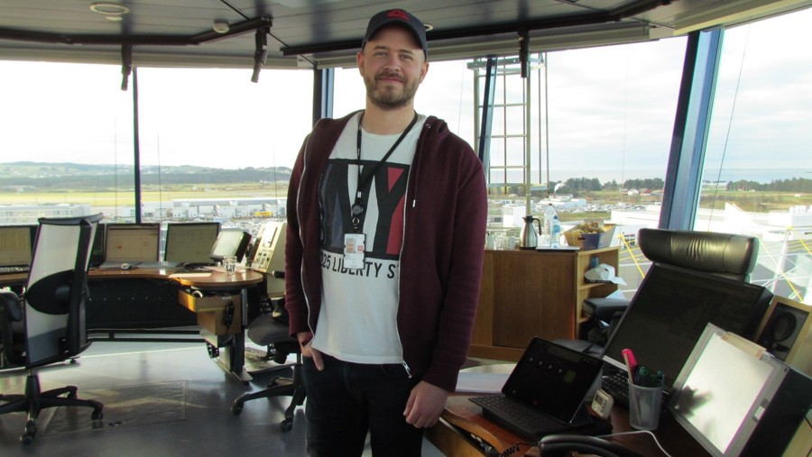 Man in control tower overlooking airport, computer screens and equipment visible in the background.