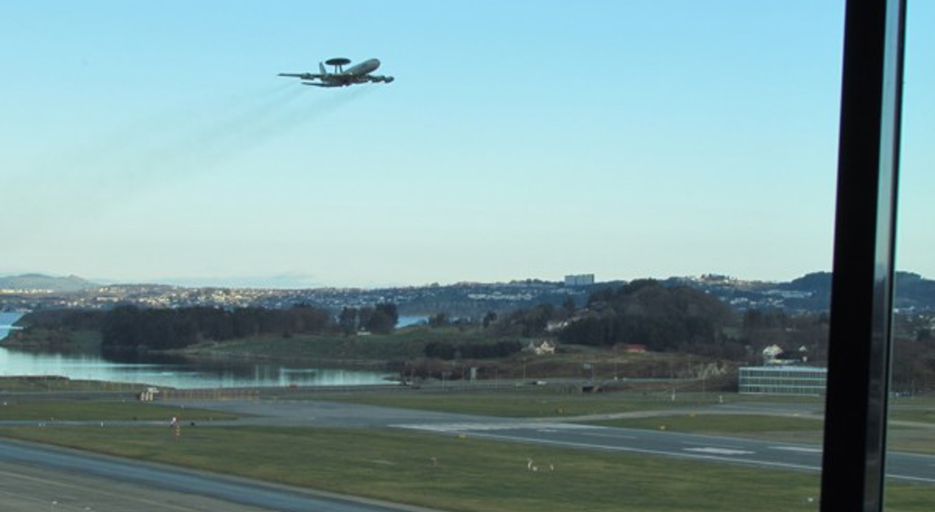 AWACS-overvåkingsfly tar av fra en flyplass med utsikt over landskap og en by i bakgrunnen i klar blå himmel.