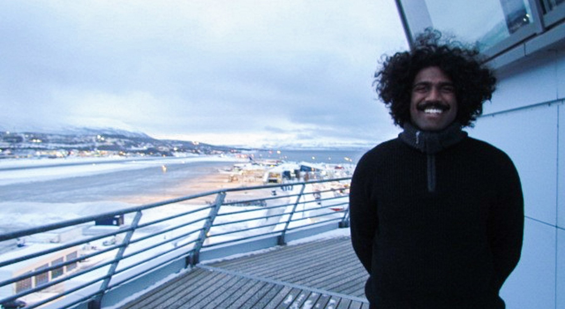 Mann som står på en balkong med utsikt over en snødekket by og vann i bakgrunnen, under en overskyet himmel.