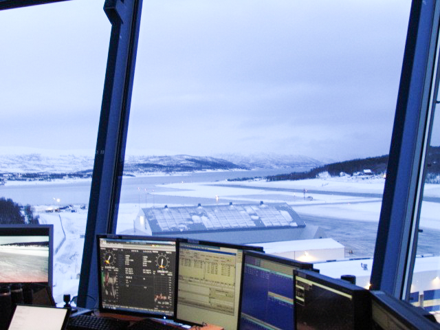 View from the control tower towards a snow-covered airport and mountainous landscape in Norway, with several computer screens in the foreground.